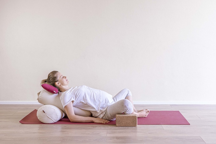 Supta Baddha Konasana A young pregnant woman lies on a yoga cushion and bolster in Reclined Wide Angle pose to alleviate constipation.